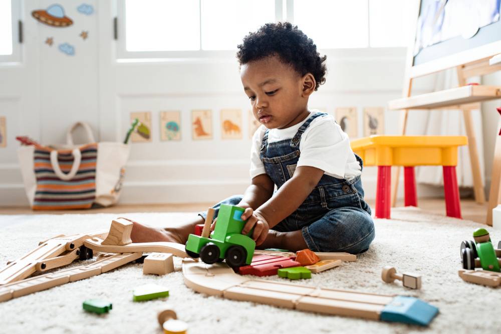 menino preto, com idade em torno de 3 anos brincando com carrinho e peças de madeira no chão da sala de brinquedos