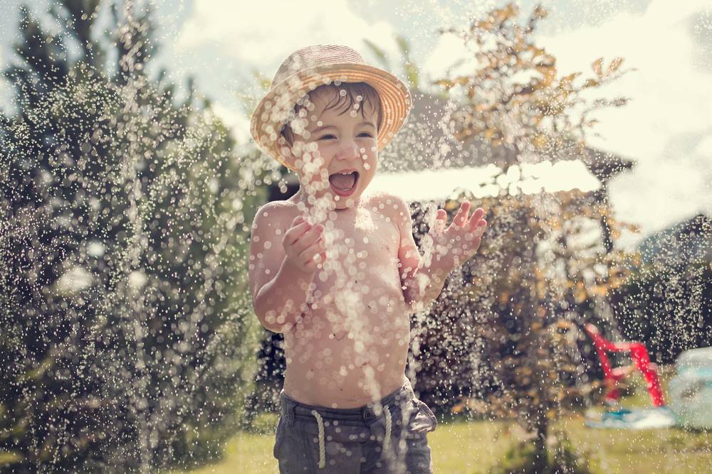 garoto branco com chapéu de palha tomando banho de água do irrigador de jardim 