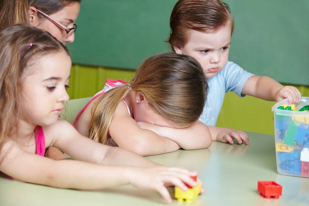 Menina branca com idade em torno de 4 anos brincando com legos em uma mesa, uma menina branca do lado dela com a cabeça deitada sobre os braços chorando e um menino branco com idade em torno de 2 anos com expressão de raiva ao lado 