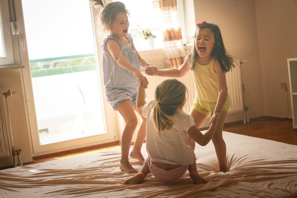 3 meninas brancas com idade entre 6 e 8 anos pulando na cama brincando e se divertindo de mãos dadas. 