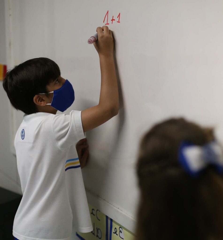 menino branco com idade em torno de 10 anos fazendo calculo em quadro branco com pincel vermelho 